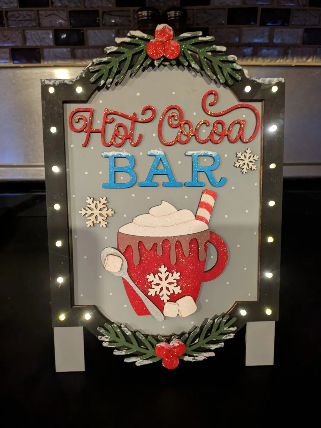 A wooden sign with lights around the border. It says 'Hot Cocoa Bar', and it has a hot cococa icon beneath that.