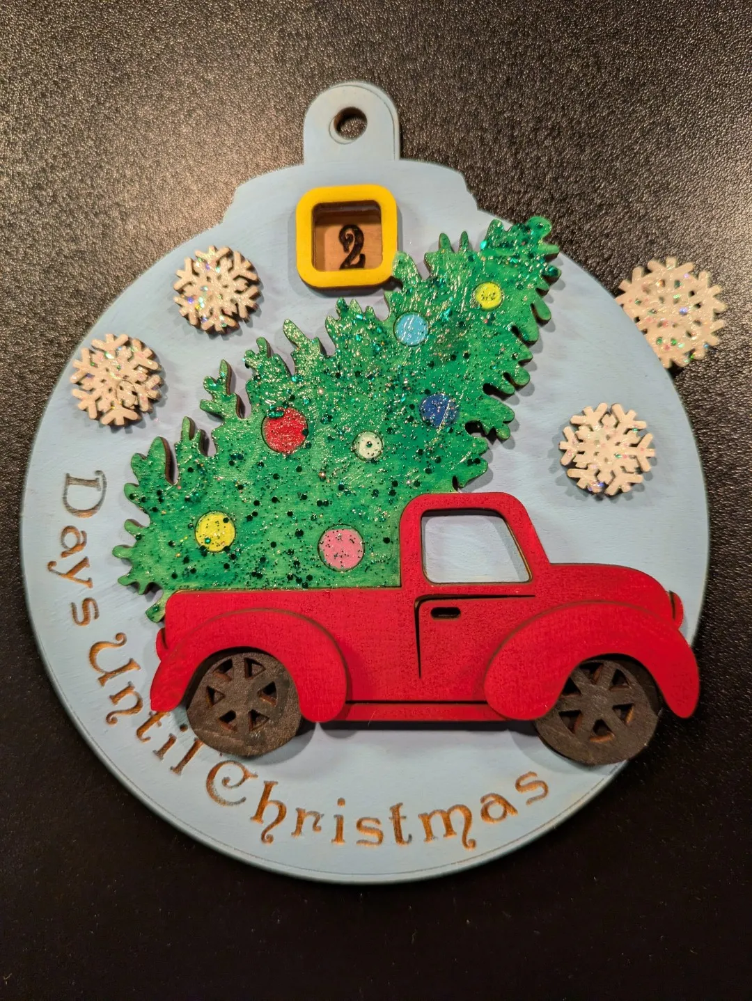 A circular wood ornament with a truck with a christmas tree. It says at the bottom, 'Days Until Christmas'. A small window at the top of the ornament displays the number '2'.