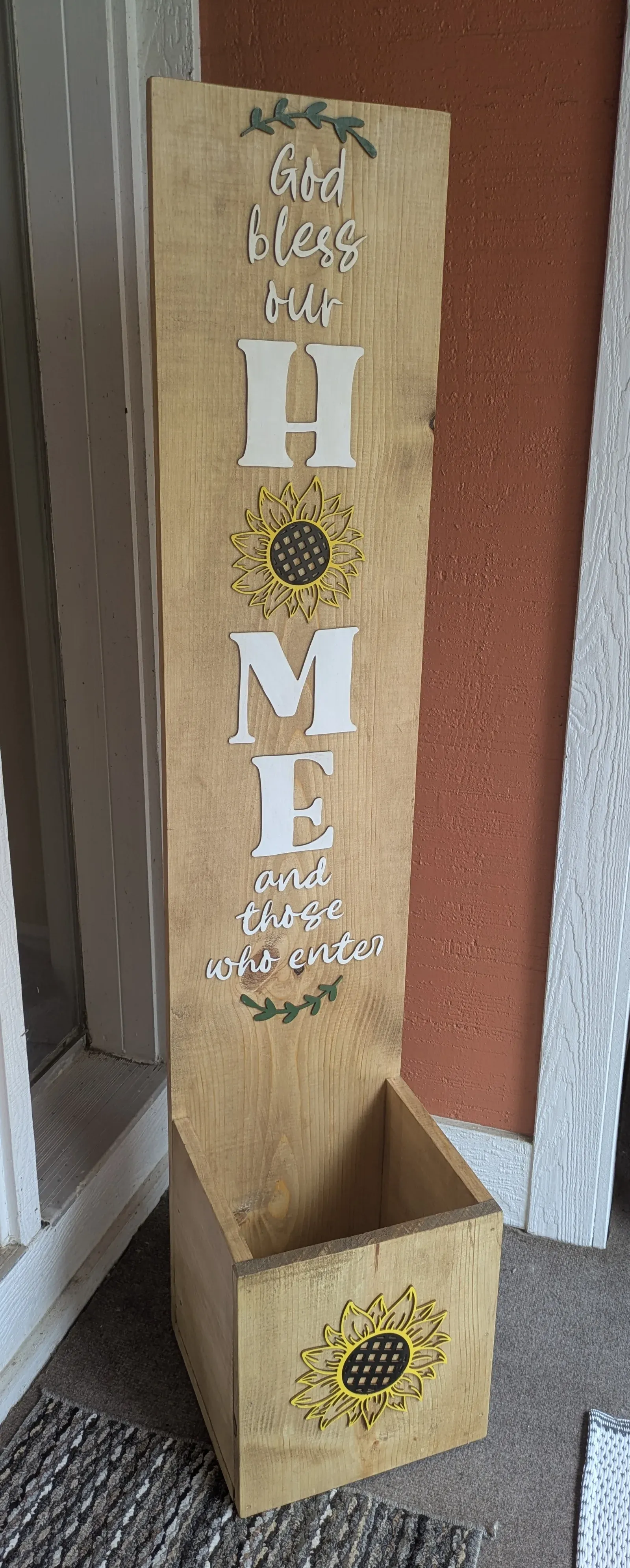 A tall light-colored wooden post with 3D layered white letters spelling out HOME with a sunflower in place of the O, surrounded by the words God bless our HOME and those who enter. A wooden planter box with a sunflower design sits at the base.