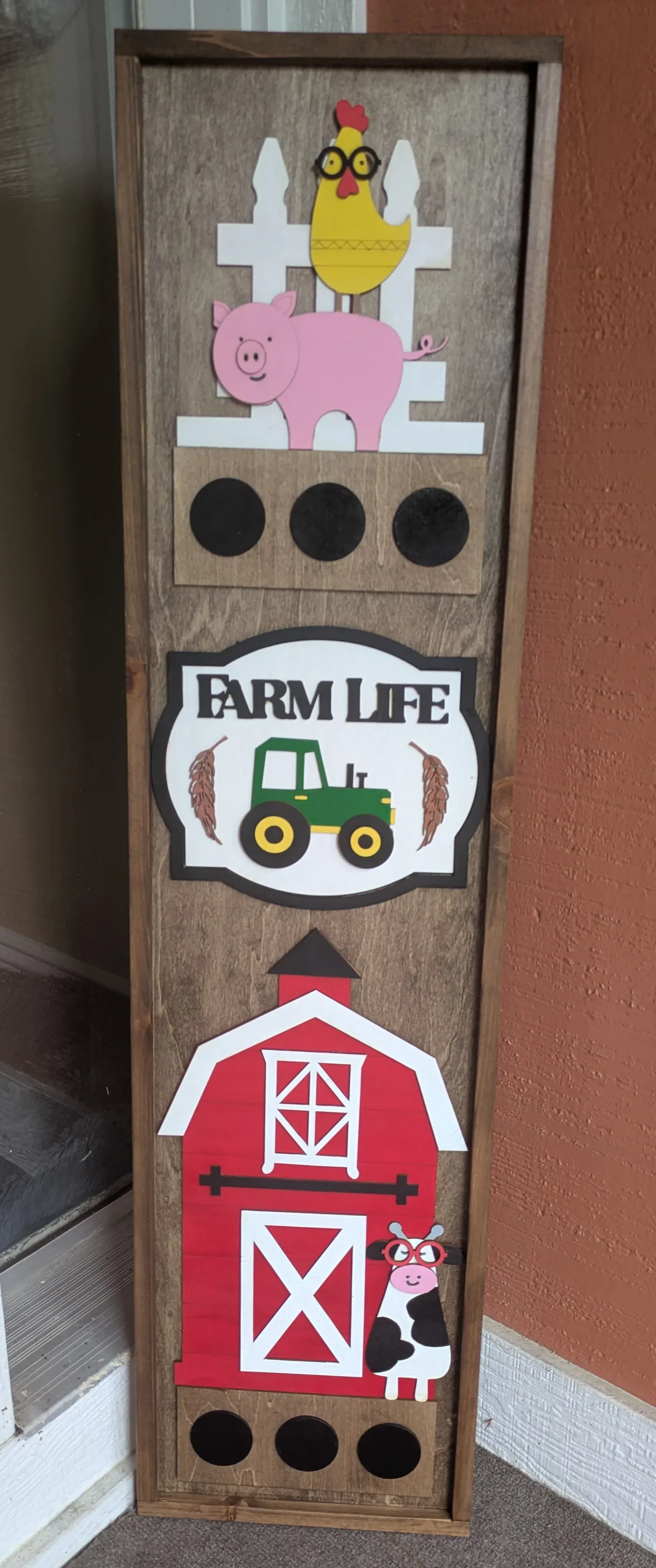 A tall vertical wooden sign leaning against a wall. It features colorful 3D farm-themed decorations: a chicken and pig above a white fence at the top, a 'Farm Life' banner with a green tractor in the middle, and a red barn with a cow peeking out at the bottom.