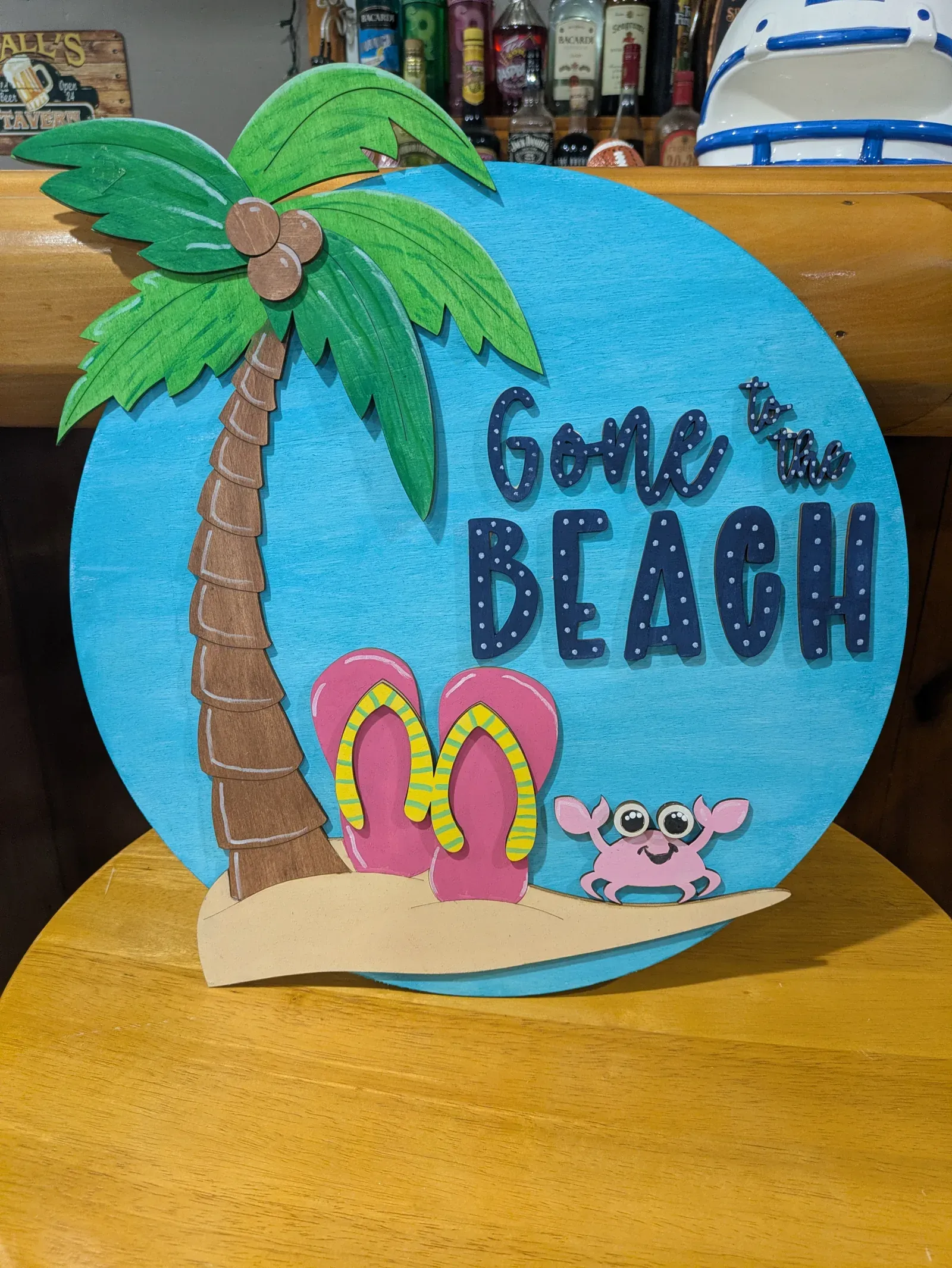 A round sign with a bright blue background depicting a palm tree, pink flip flops, a pink crab, and the words 'Gone to the Beach' in navy script.