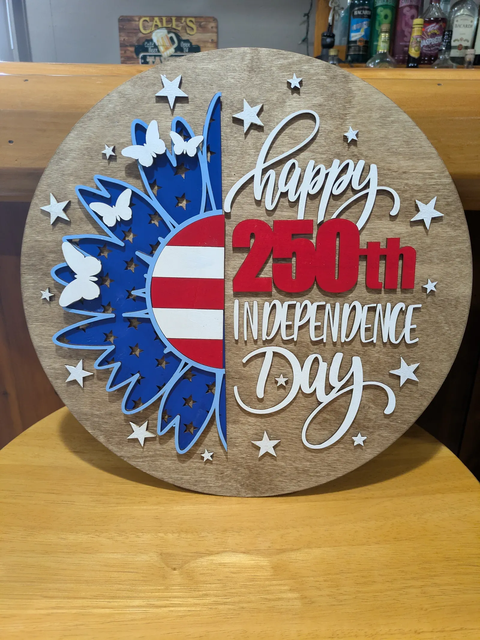 A round wooden sign celebrating America's 250th Independence Day, featuring a sunflower in red, white, and blue with butterflies and stars, and text reading 'Happy 250th Independence Day'.