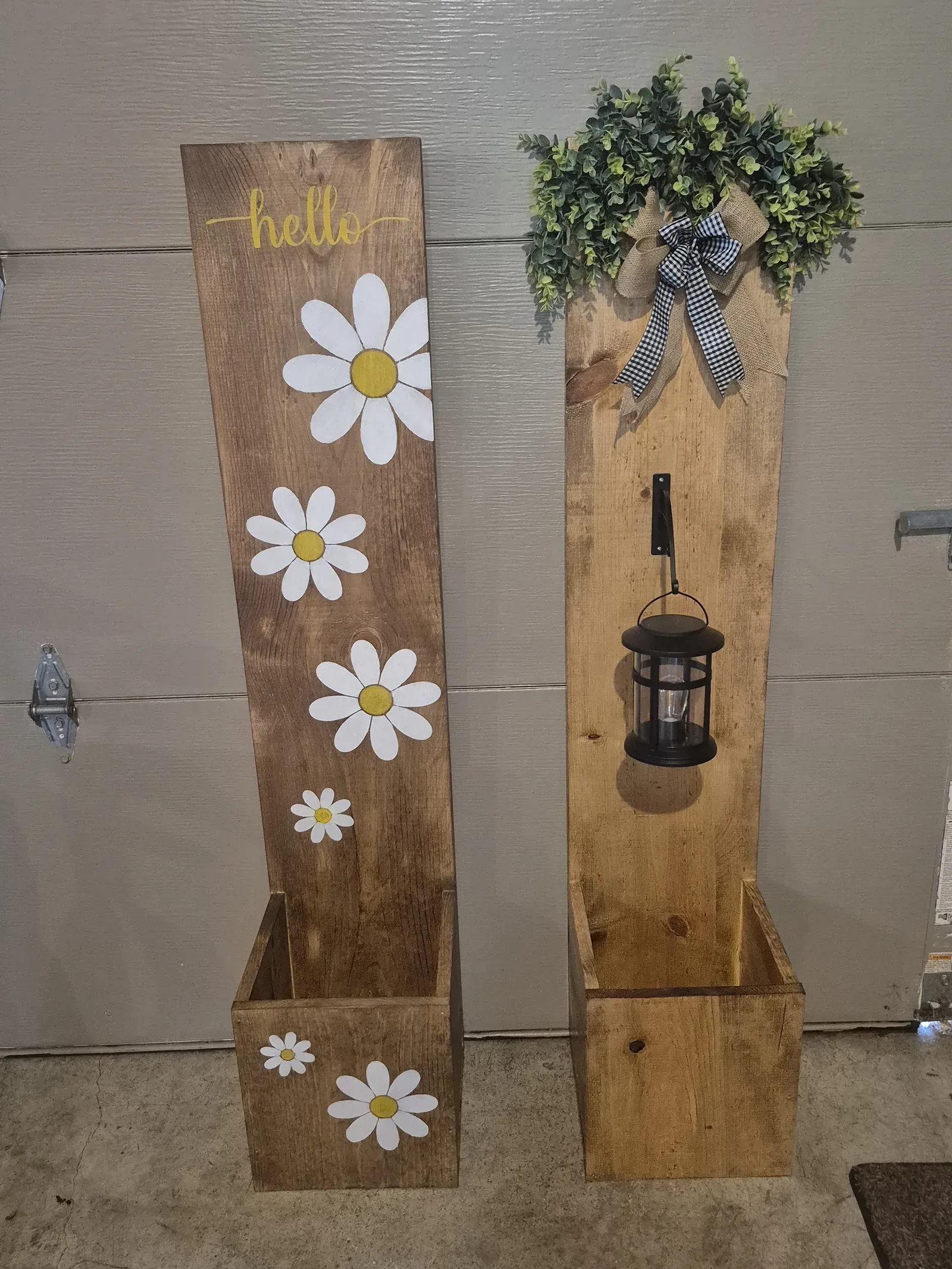 Two rustic stained wooden planter posts leaning against a wall side by side. The front post is painted with white daisy flowers and the word 'hello' in yellow script. The post behind it is topped with a hanging black metal lantern and decorated with a green eucalyptus wreath and navy gingham ribbon.
