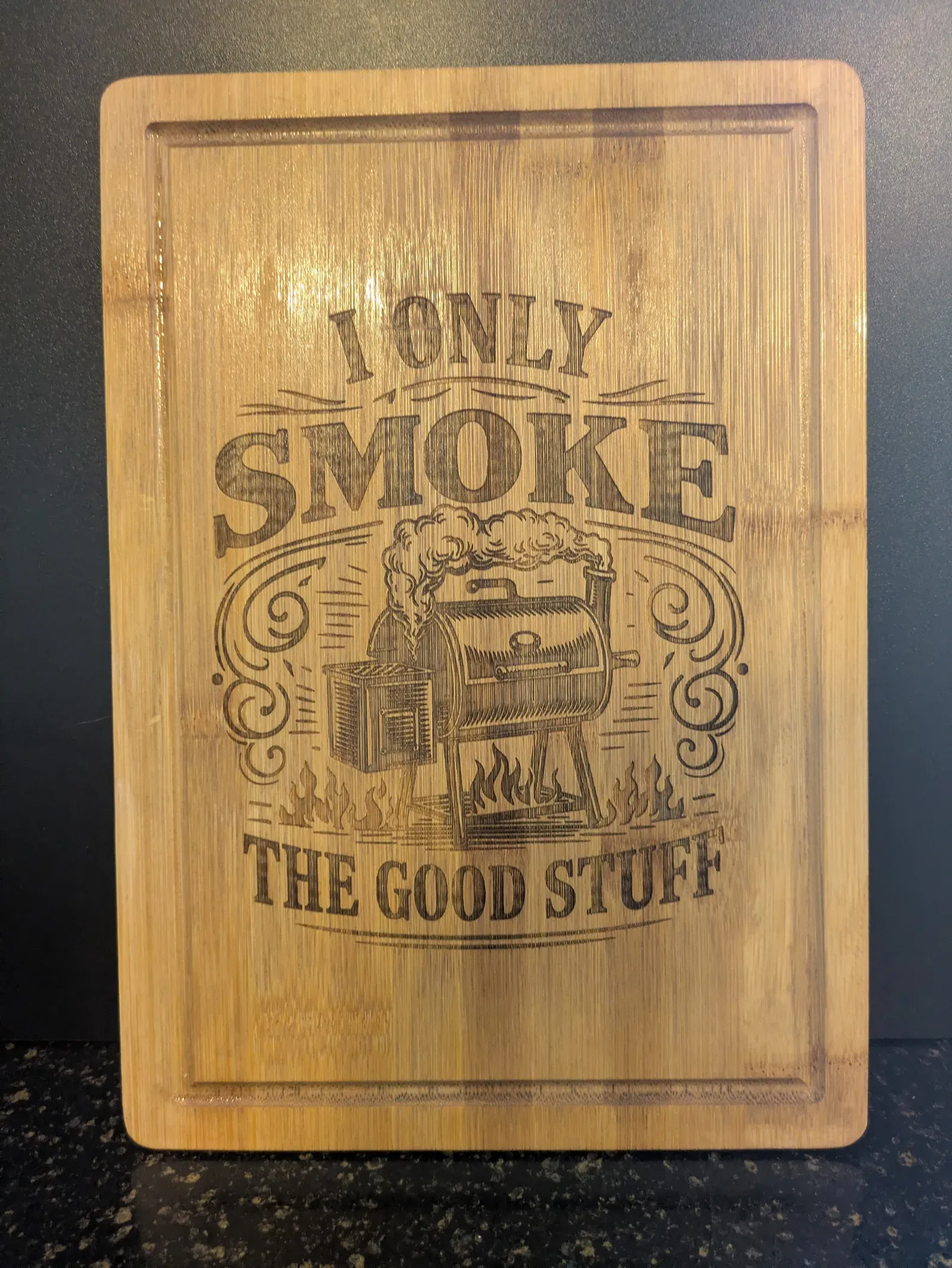 A bamboo cutting board laser-engraved with 'I Only Smoke the Good Stuff' in bold lettering surrounding an illustration of a BBQ smoker grill with decorative flourishes.