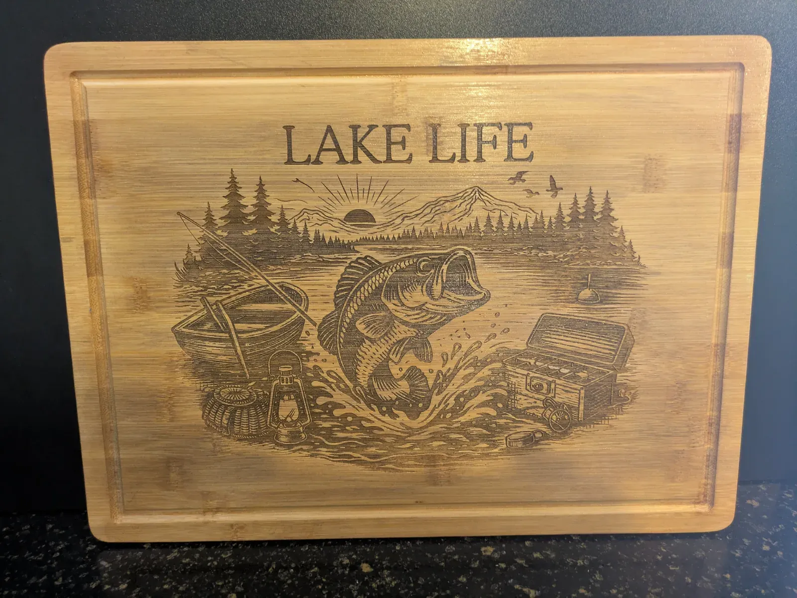 A large bamboo cutting board laser-engraved with a detailed Lake Life scene: a bass jumping out of the water surrounded by pine trees, a mountain sunset, a lantern, and a tackle box.