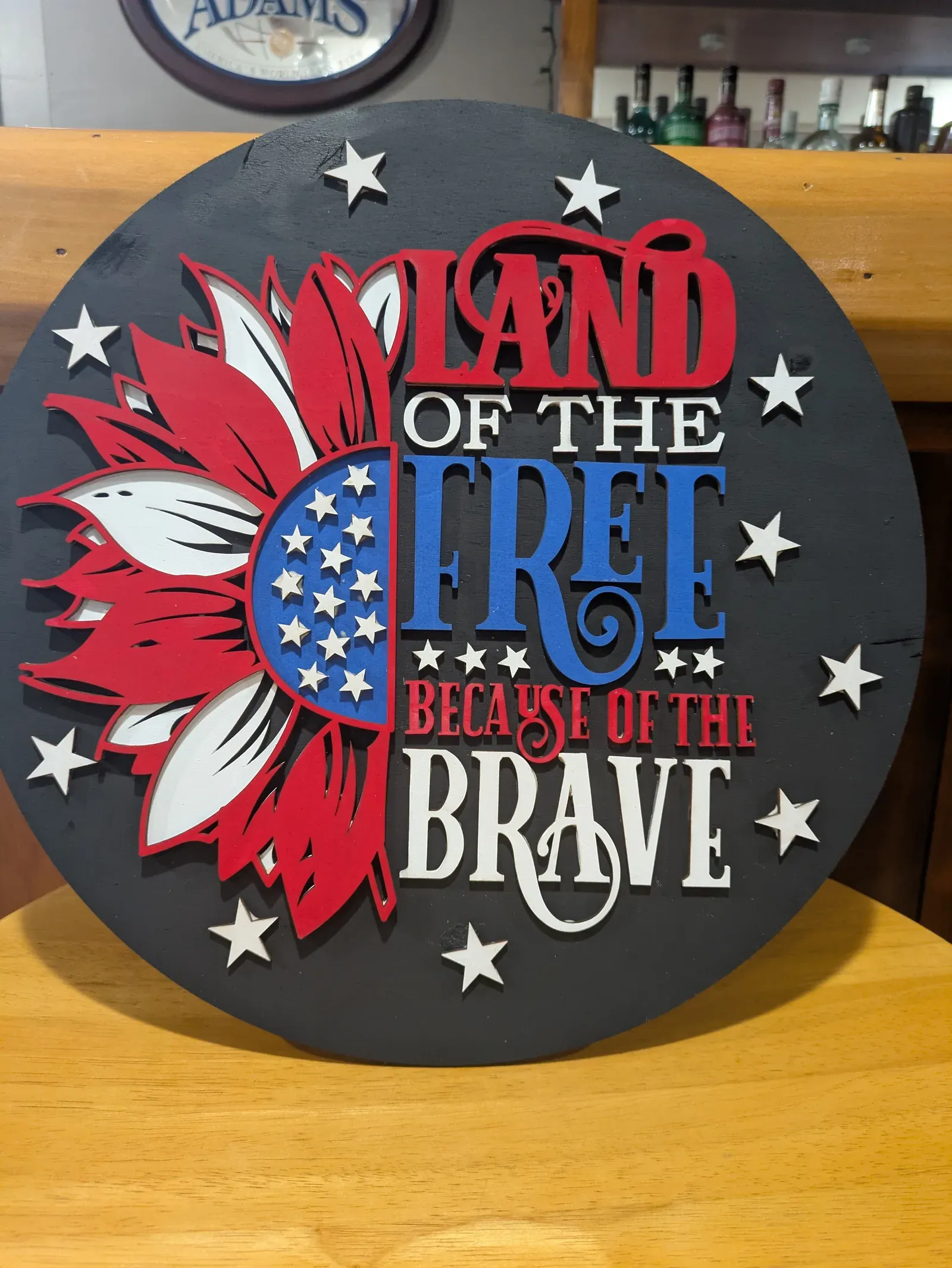 A large round sign with a dark background featuring a red, white, and blue sunflower on the left and bold text reading 'Land of the Free Because of the Brave' surrounded by white stars.
