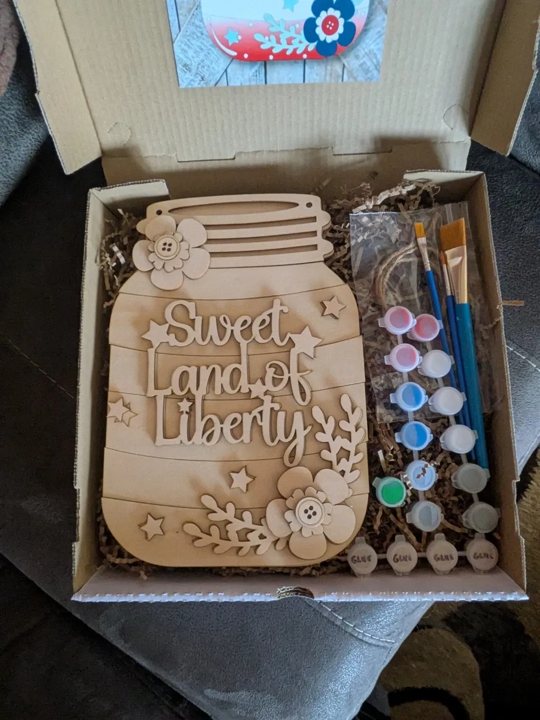 A DIY kit in a box with a wood piece engraved to look like a jar reading 'Sweet Land of Liberty' with flowers, plus paint, brushes, and packing materials.