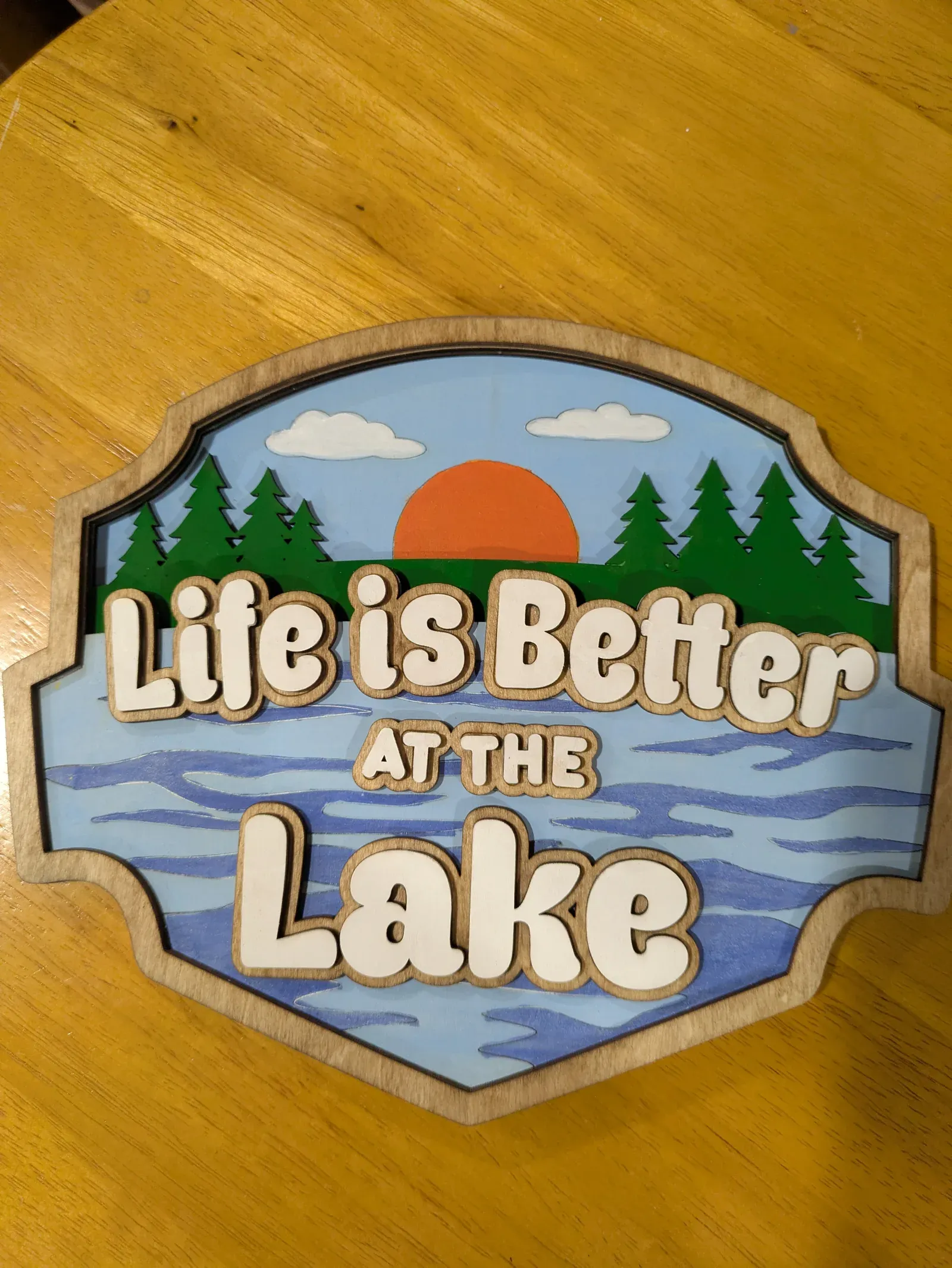 A shaped wooden sign with a scenic lake design: pine trees, a bright orange sunset, clouds, and water with the words 'Life is Better at the Lake' in white lettering.