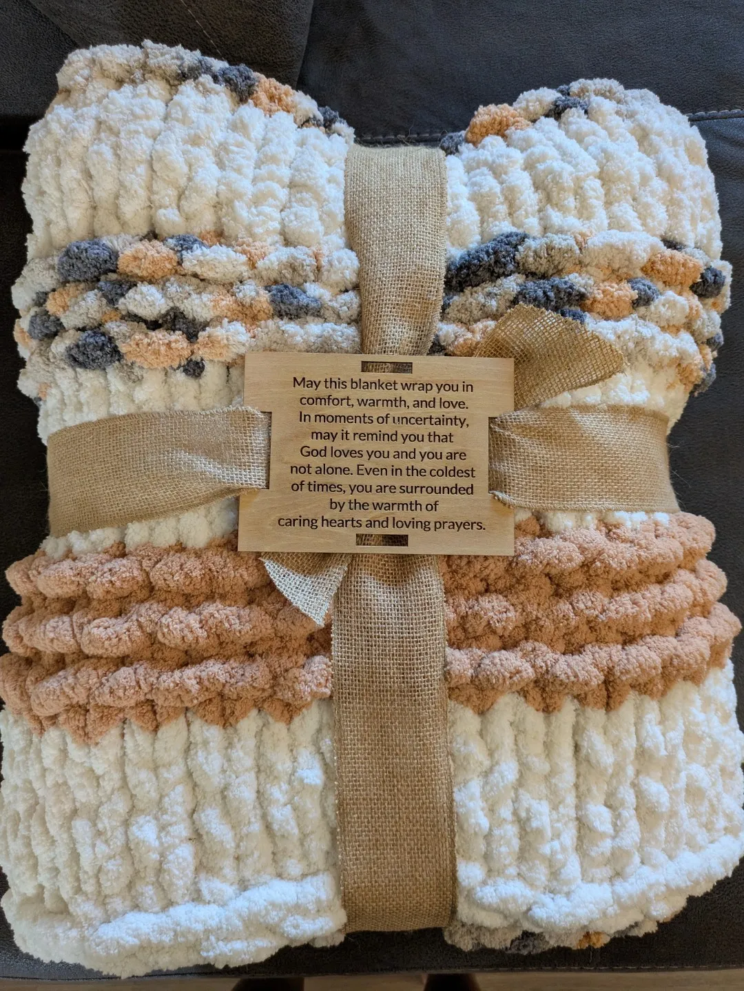 A blanket made from white, orange and gray fabric. It is folded and wrapped, held together by a wooden plaque with text.