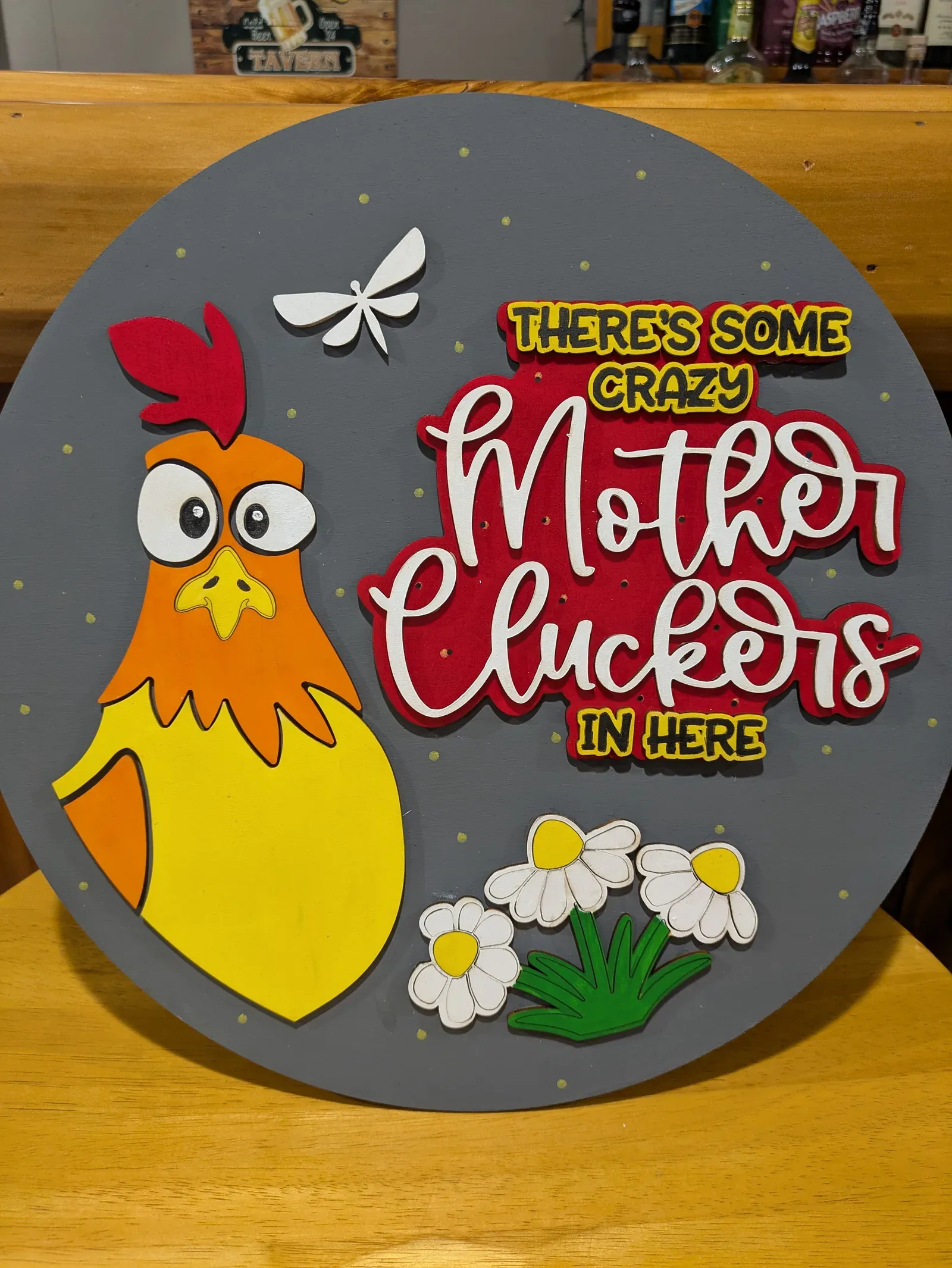 A round sign with a gray background featuring a cartoon yellow chicken, a white dragonfly, white daisy flowers, and the humorous text 'There's Some Crazy Mother Cluckers in Here' in red and white lettering.