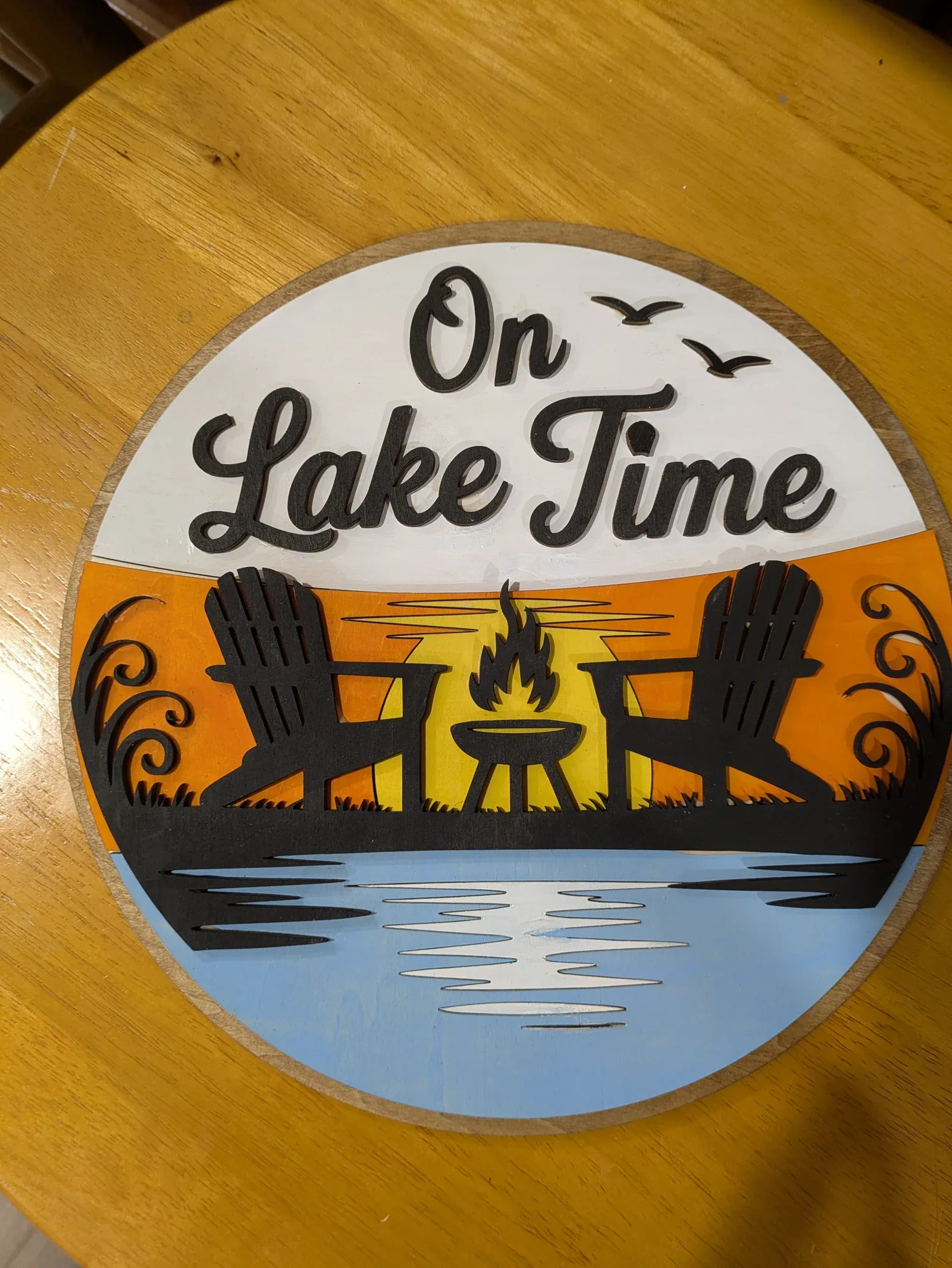 A round sign featuring a peaceful lake scene at sunset with two Adirondack chairs, a campfire, and cattails reflected in the water, with 'On Lake Time' written in bold script at the top.