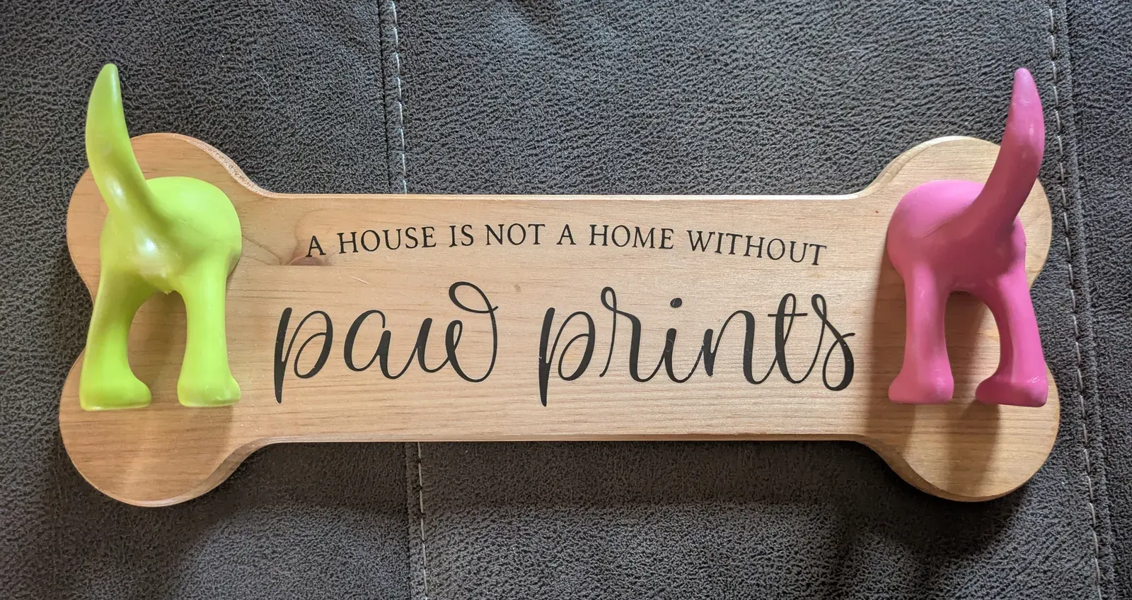 A bone-shaped wooden sign with 'A House Is Not a Home Without Paw Prints' laser-engraved in the center. A bright green 3D dog butt figure is mounted on the left end and a pink one on the right, each serving as a hook for hanging pet collars or leashes.