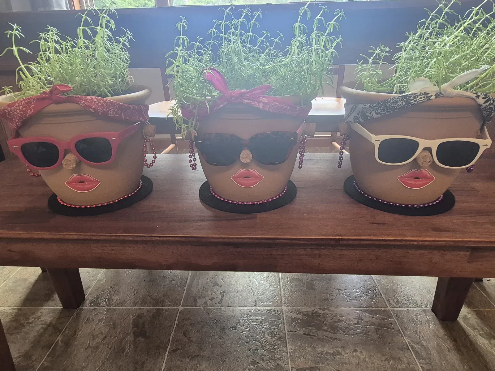 Three terracotta pots sitting on a wooden bench, each decorated to look like a sassy woman — pink sunglasses, a red bandana, a sparkly headband — with red lips, earrings, bead necklaces, and lush green herbs growing from the tops.
