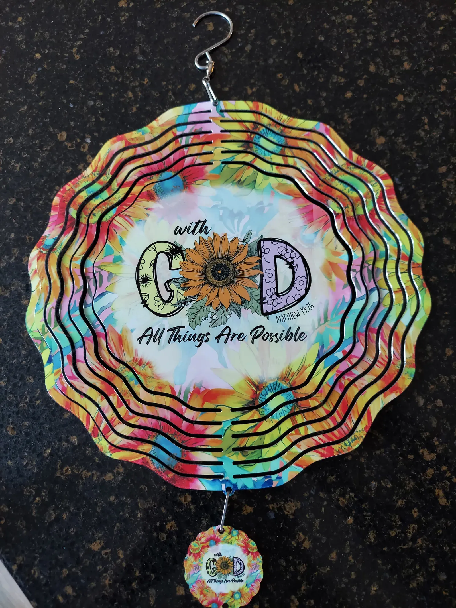 A flat circular metal wind spinner with a tie-dye rainbow background featuring a sunflower in a pot and the text 'with God All Things Are Possible'. A small matching charm hangs below.
