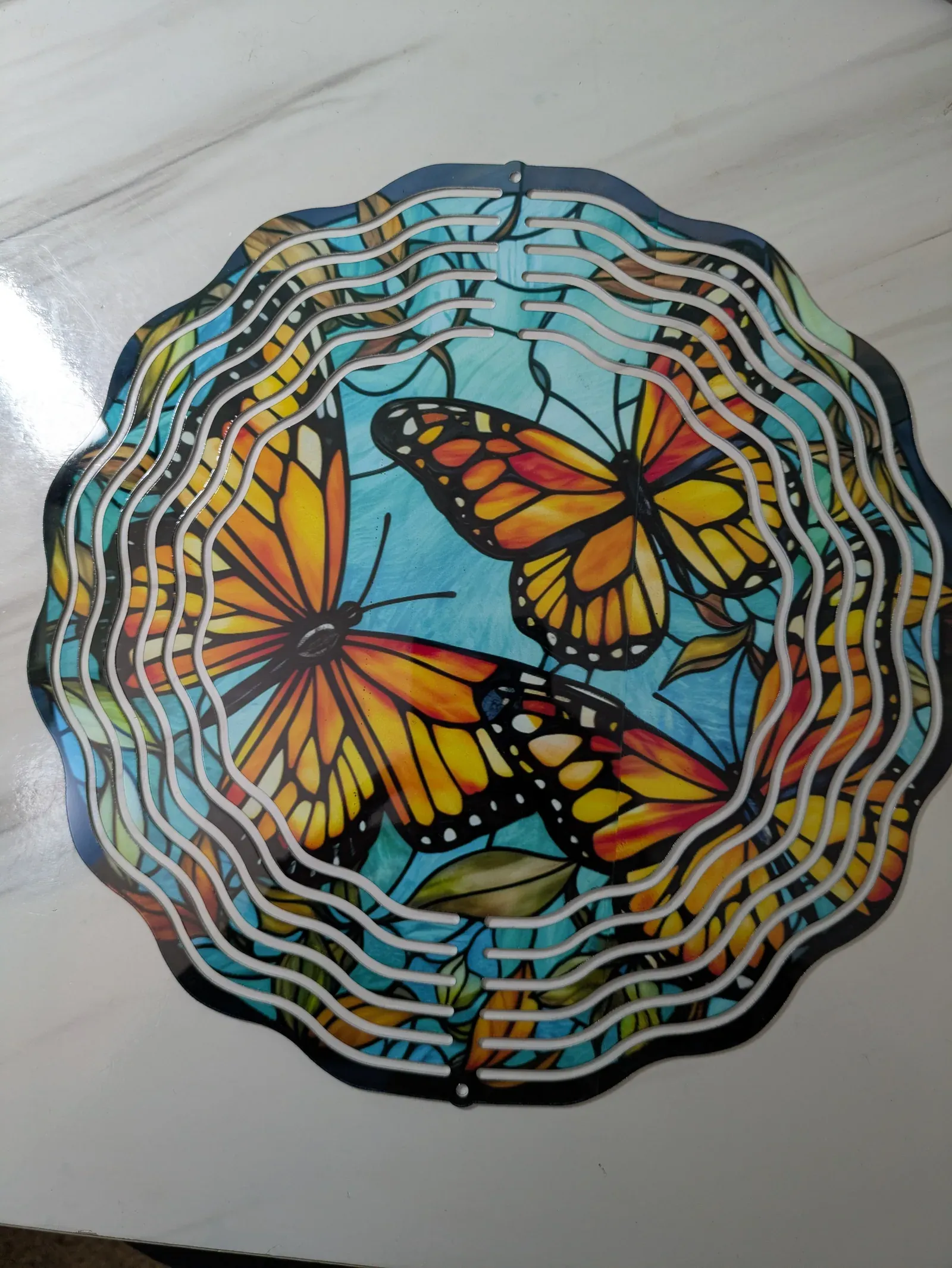 A flat circular metal wind spinner with a colorful design of orange and yellow monarch butterflies on a blue background, surrounded by concentric wavy rings in iridescent colors.