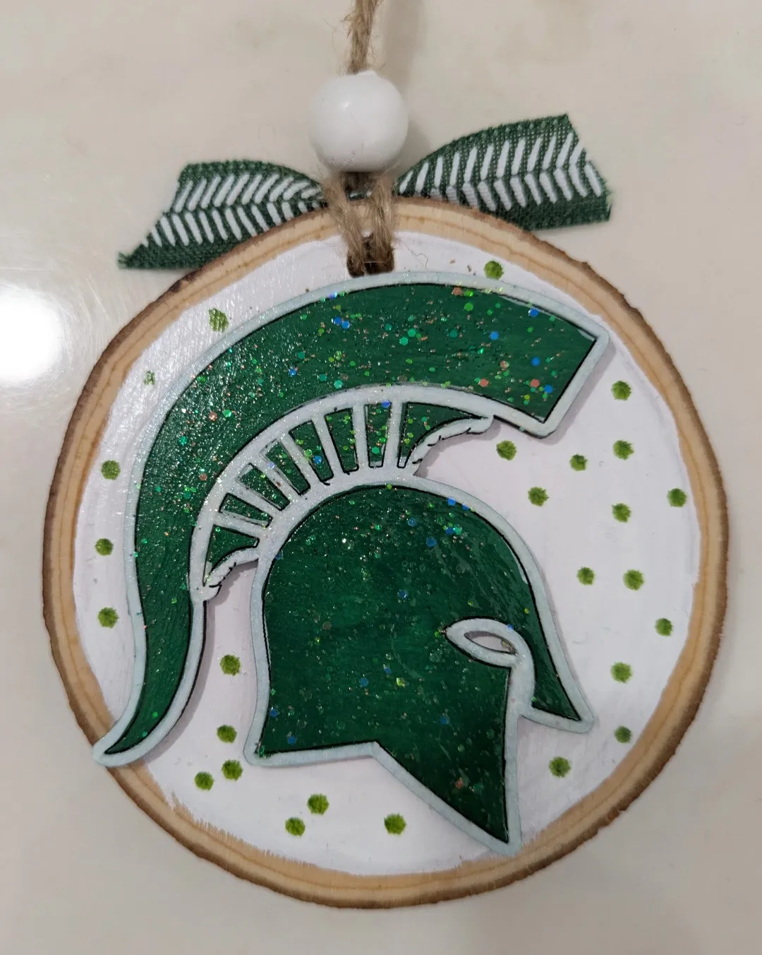 A wooden ornament. It is a white circle with green dots and a green Michigan State Spartans head jutting out from the ornament.