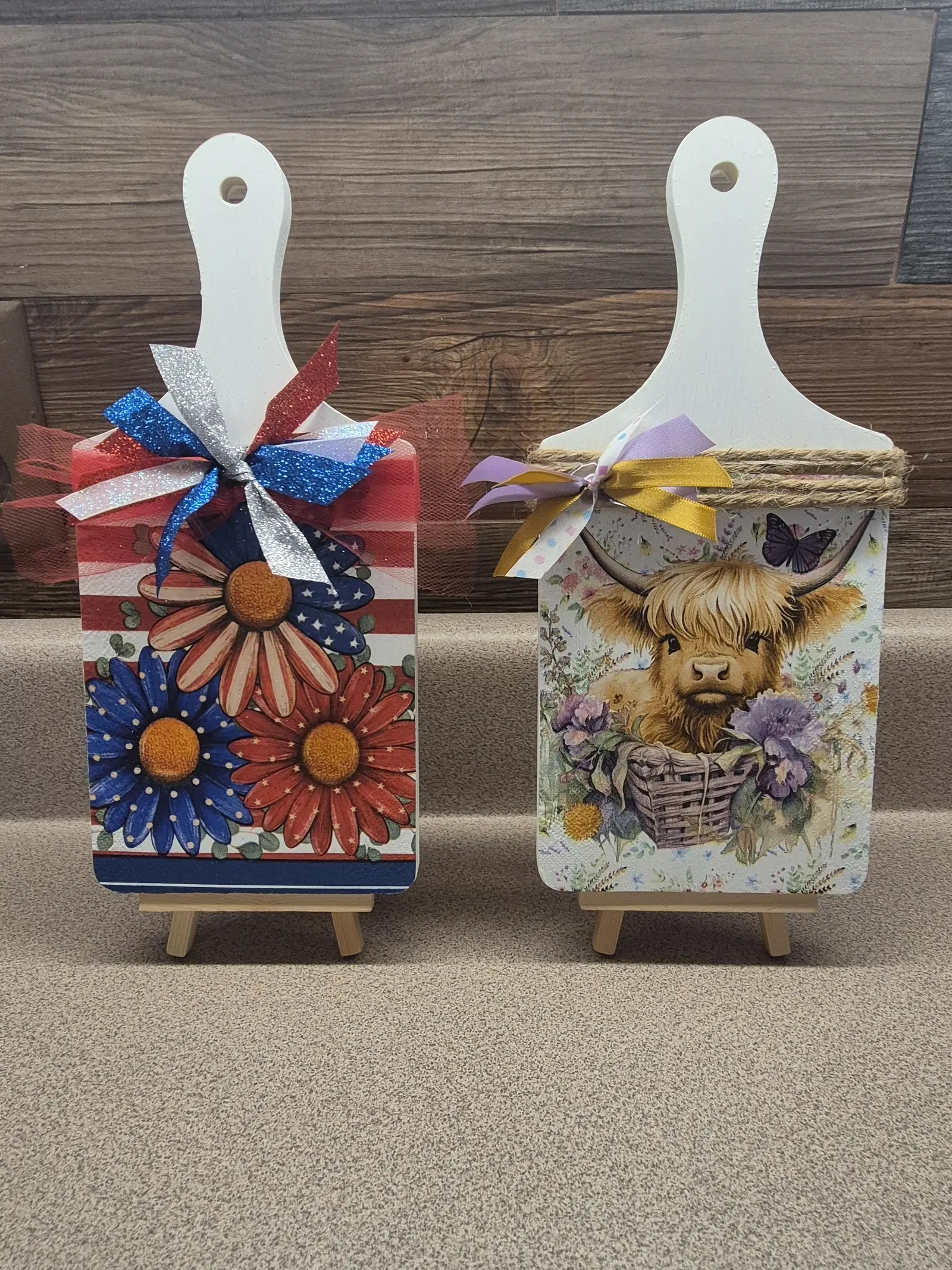 Two small decorative wooden shelf sitters propped on wooden easel stands. The left one has a highland cow amid purple and yellow wildflowers with a lavender ribbon. The right one features bold red, white, and blue daisy flowers in a patriotic design with a red, white, and blue glitter ribbon.