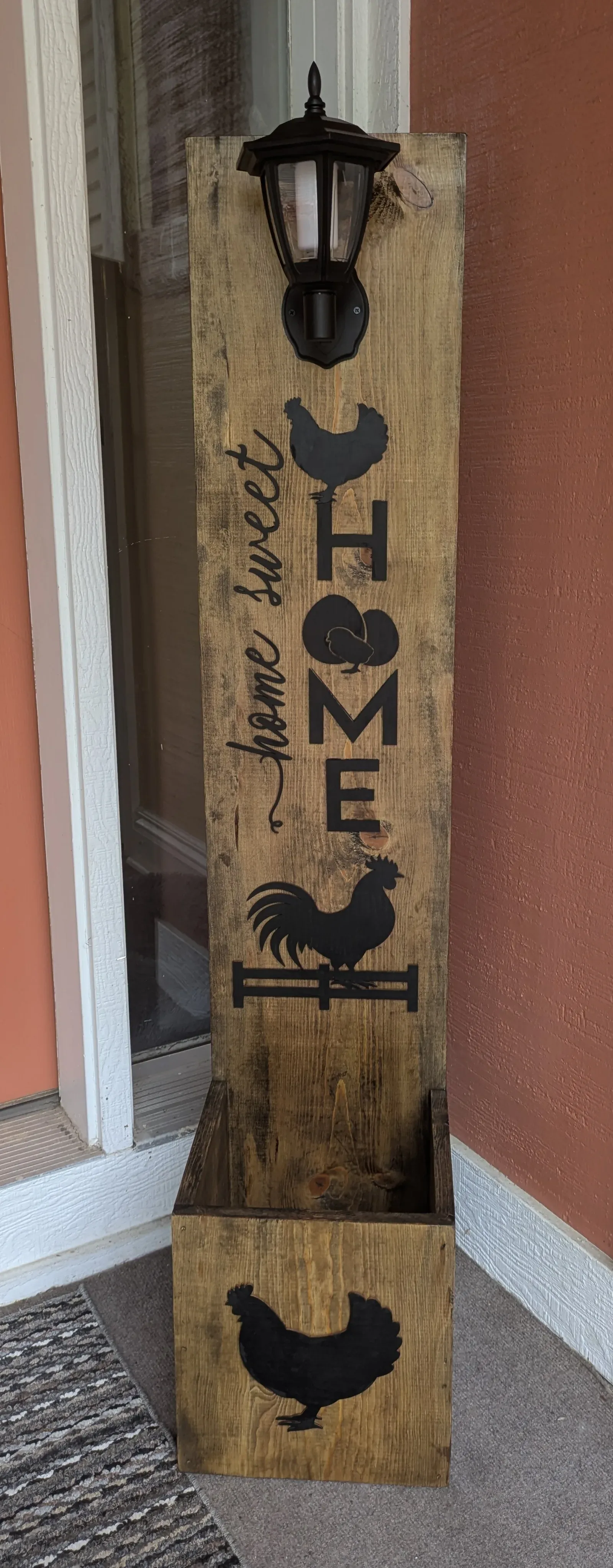 A tall rustic wooden post leaning against a front door, painted dark brown with 'home sweet home' lettered in script and illustrated farm animals including chickens and a rooster. A solar-powered black lantern sits on top. A matching wooden planter box with a chicken silhouette sits at the base.