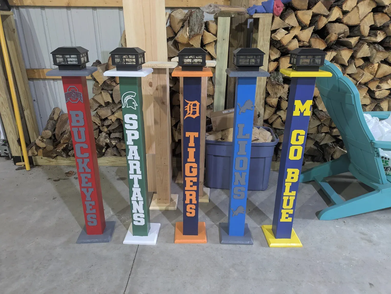 Five tall wooden lantern posts lined up side by side, each painted in a different team's colors — red for Ohio State Buckeyes, green for Michigan State Spartans, orange for Detroit Tigers, blue and silver for Detroit Lions, and blue and yellow for Michigan Wolverines. Each post is topped with a black solar-powered lantern.