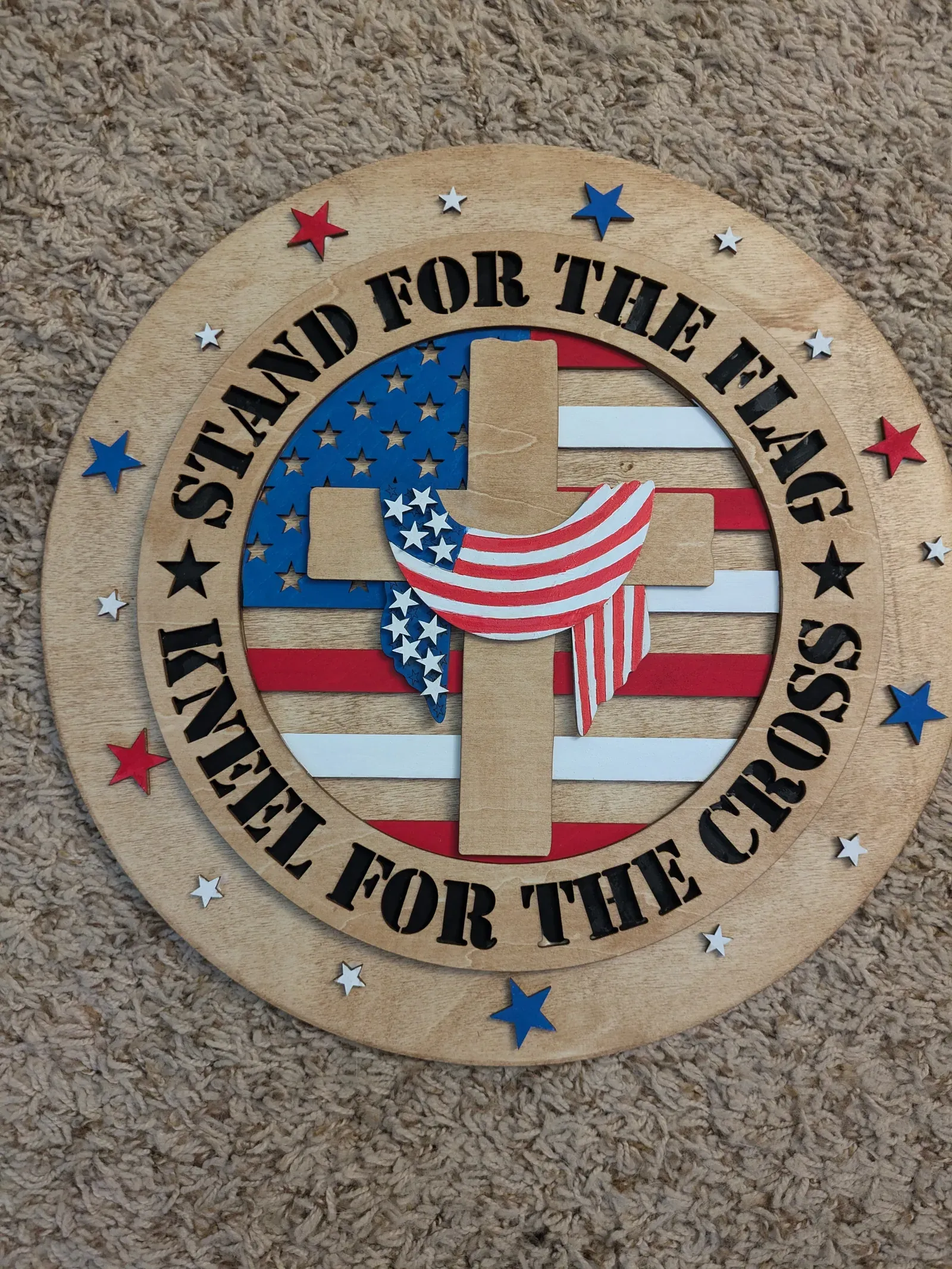 A large round wooden sign with a patriotic design: a cross draped in an American flag in the center, surrounded by red, white, and blue stars, with the words 'Stand for the Flag, Kneel for the Cross' around the border.