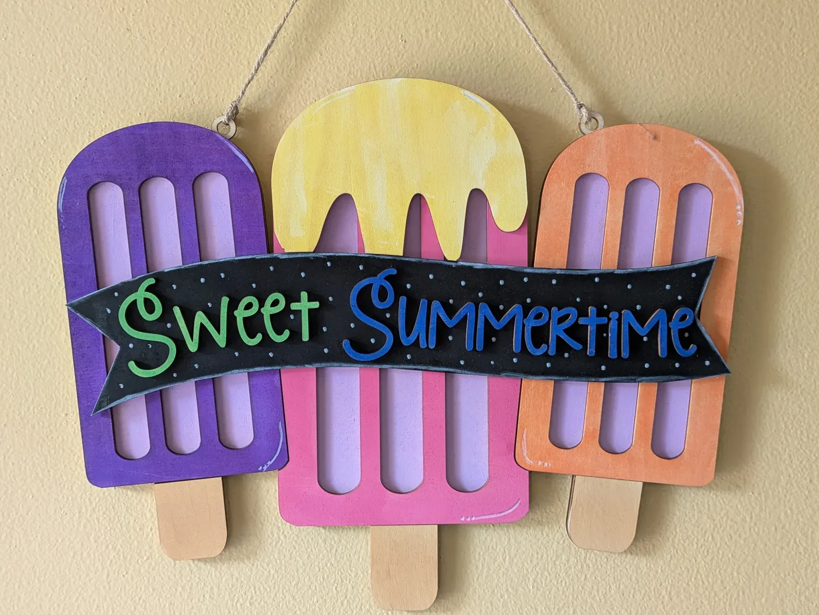 A hanging wooden sign made of three large popsicle shapes in purple, yellow, and orange with popsicle sticks at the bottom. A black banner across the middle reads 'Sweet Summertime' in green and blue lettering. The yellow center popsicle has a dripping top.