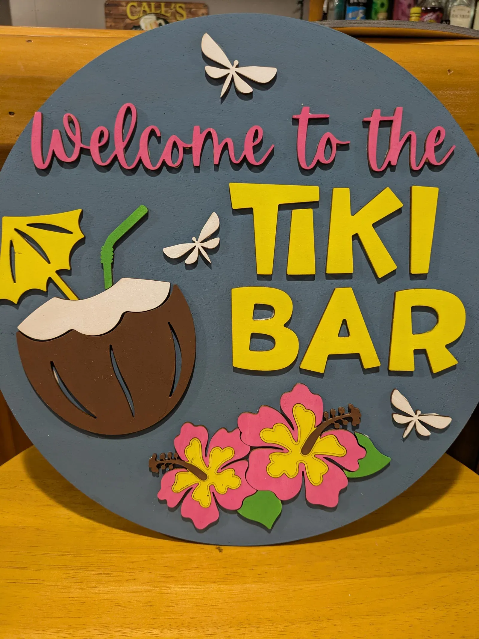 A round sign with a dark blue-gray background featuring a tropical coconut drink, pink hibiscus flowers, white dragonflies, and bold yellow text reading 'Welcome to the Tiki Bar'.