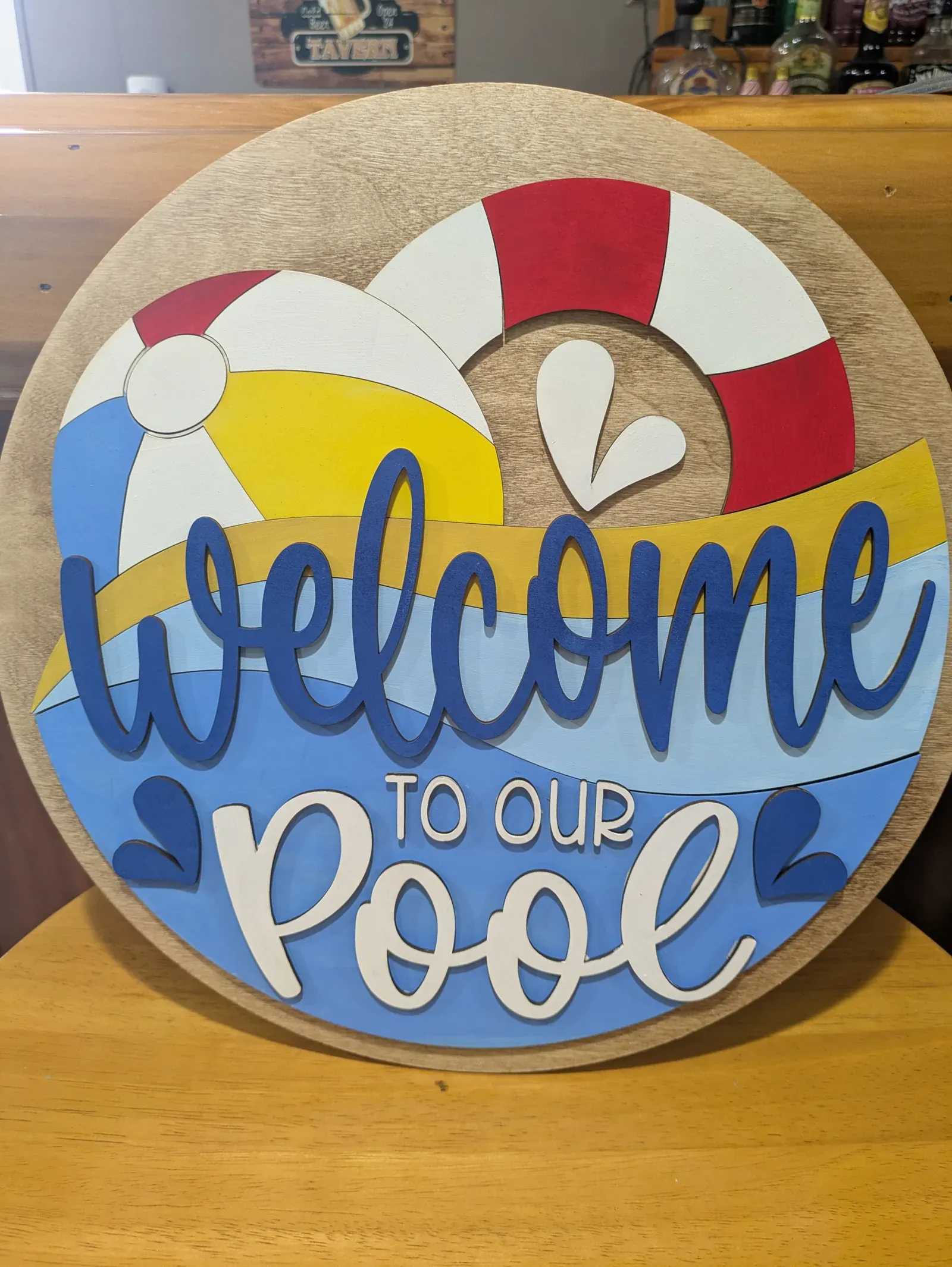 A round sign with a natural wood border and a summer pool scene: blue waves, a beach ball, a red and white life preserver, and the words 'Welcome to Our Pool' in blue script.