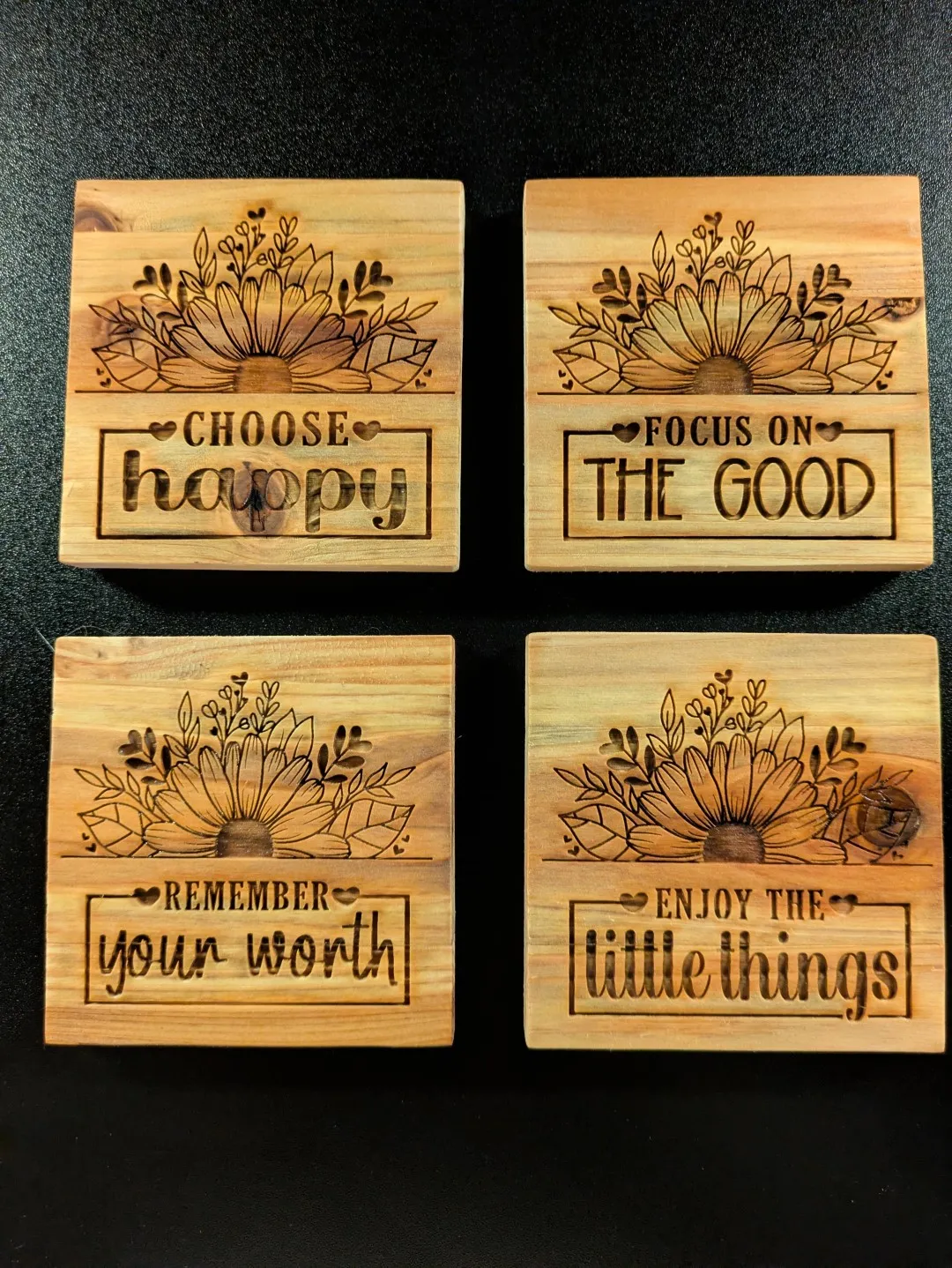 A set of 4 wooden coasters. Each one has a sunflower image surrounded by some smaller plants. Each one also has a message, which are: 'Choose happy', 'Focus on the good', 'remember your worth', and 'Enjoy the little things'.