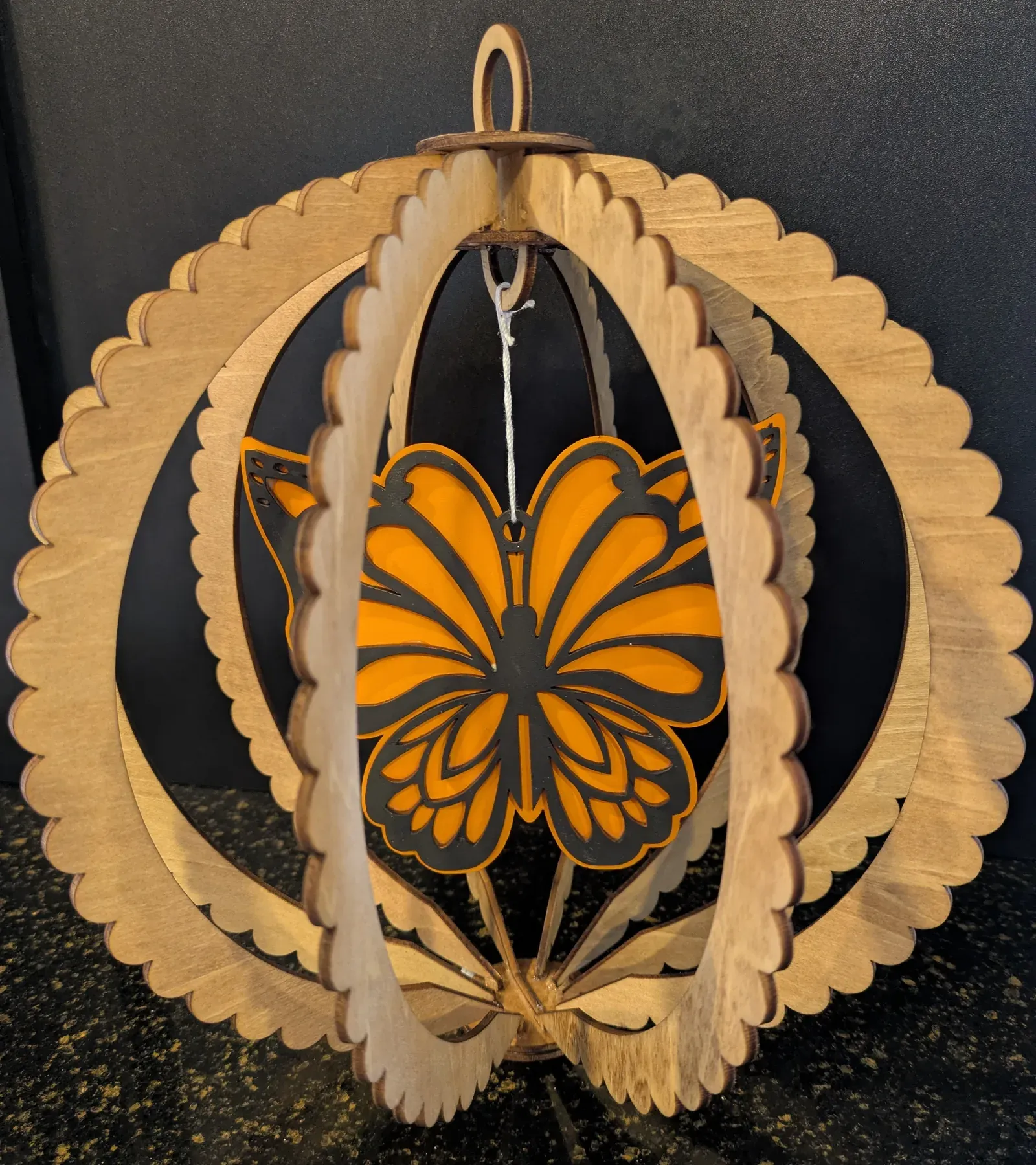 A 3D globe-shaped wooden wind spinner with an orange and black butterfly suspended in the center, surrounded by layered wooden rings with scalloped edges.