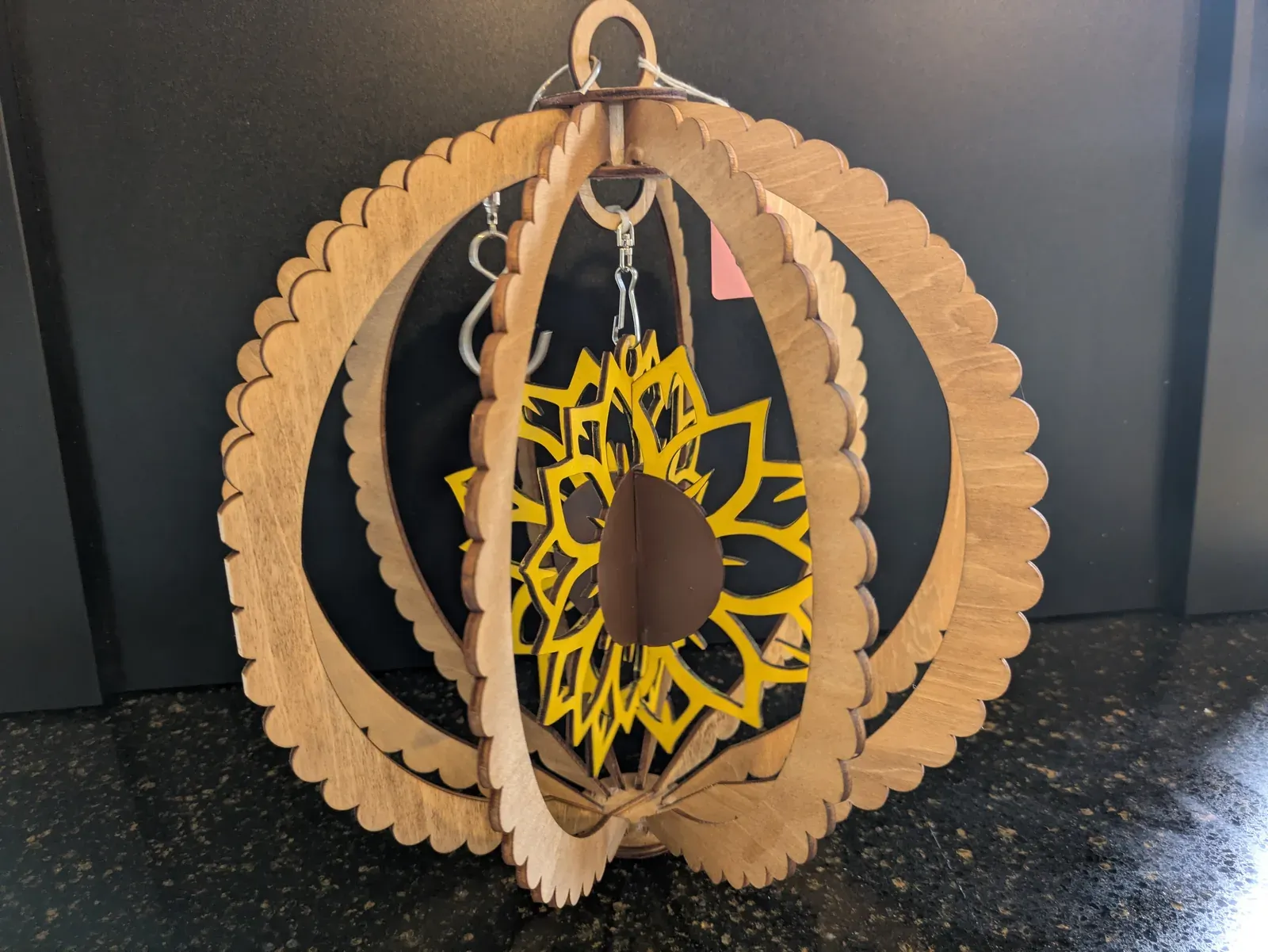 A 3D globe-shaped wooden wind spinner with a large yellow sunflower suspended in the center, surrounded by layered wooden rings with scalloped edges. A hanging loop sits at the top.