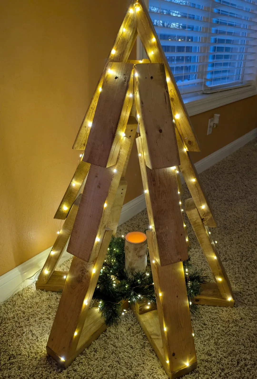 A christmas tree structure created out of pieces of unpainted wood. An LED light strip adorns the sides. At the base of the tree, in the middle, is a wreath with more lights and a candle.